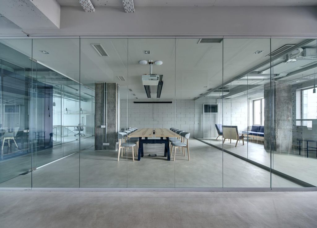 Office in a loft style with white brick walls and concrete columns. There is a meeting zone with a large wooden table with gray chairs and glass partitions. Above the table there is a projector.